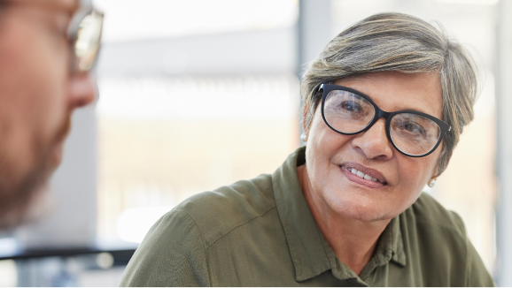 A mature woman wearing glasses looks over to her coworker as he explains something.