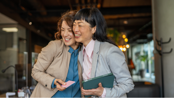 Two smiling women in professional attire give each other a side hug.