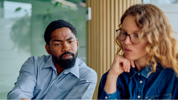 A man seated next to his female coworker leans in to view what she is explaining.