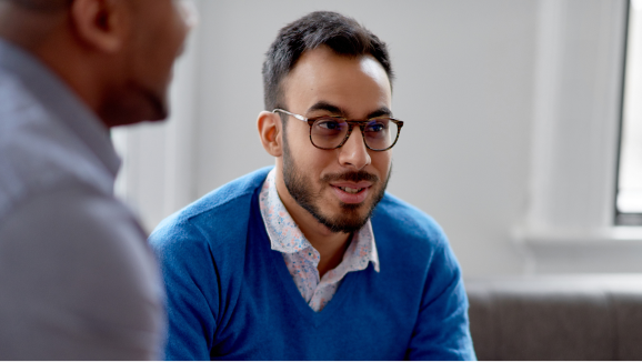 A man wearing glasses and professional attire sits while speaking to a coworker.