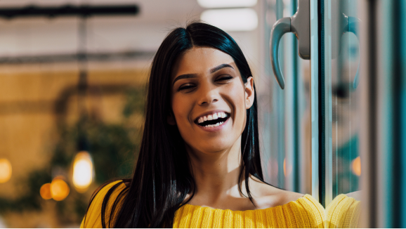 A woman with long, dark hair smiles while standing near a window.