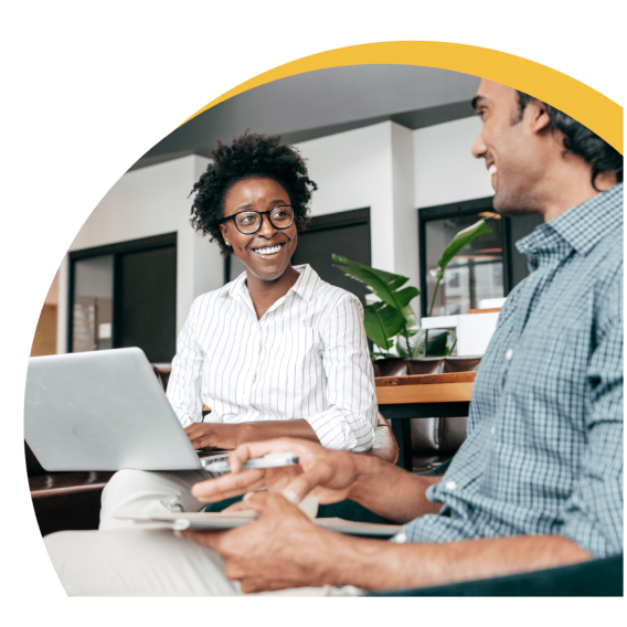 A woman with a smile working on a laptop and interacting with a coworker