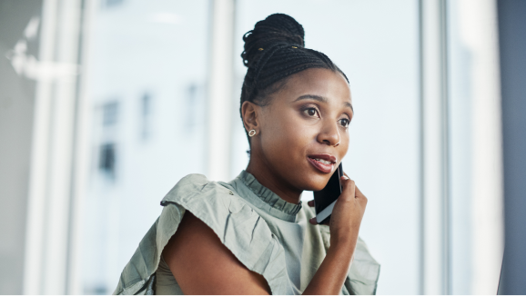 A woman wearing professional attire takes a phone call while sitting near an office window.